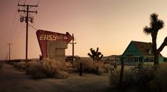 an old gas station in the desert with palm trees and telephone poles around it at sunset