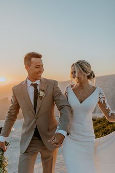 a newly married couple holding hands and walking on the beach at sunset in their wedding attire