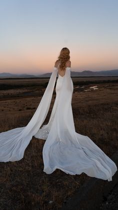 a woman in a white dress is walking through an open field with her long veil blowing in the wind