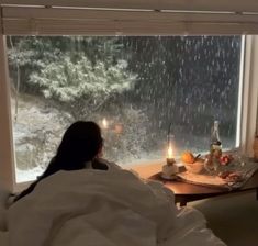 a woman sitting on a bed in front of a window looking out at the rain