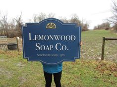 a woman holding up a sign that says lemonwood soap co in front of a field