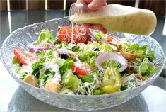 a person pouring dressing over a salad in a glass bowl on top of a table