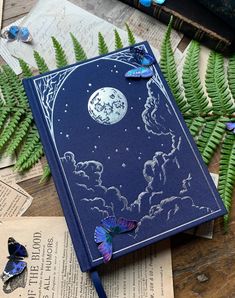 a blue book sitting on top of a wooden table next to green plants and butterflies