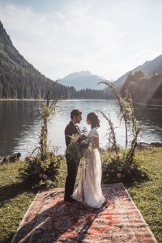 a bride and groom standing on a rug in front of a lake surrounded by greenery