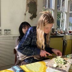 two women sitting at a table cutting up food on top of a cutting board with knifes