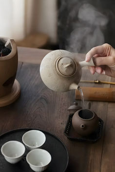 a person pouring tea into small cups on a wooden table next to two pots and a bamboo stick