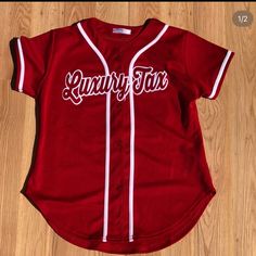 a red baseball jersey with the word kentucky on it and white stitching is laying on a wooden floor