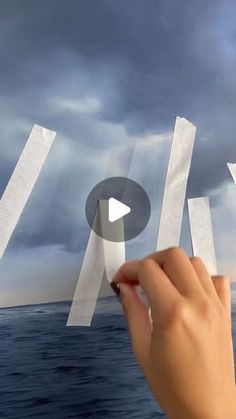 someone is holding some paper in their hand over the water and clouds above them that look like they are flying through the air