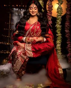 a woman in a red and gold sari sitting on a chair with flowers around her