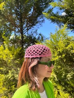 a woman wearing sunglasses and a pink crochet hat in the woods with trees behind her