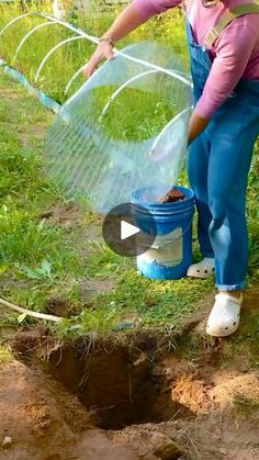 a woman in overalls and pink shirt holding a blue bucket filled with dirt next to a hole