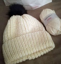 two balls of yarn and a white knitted beanie with a black pom - pom