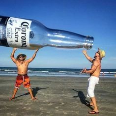 two men standing on the beach with a bottle in their hand and one man holding up his hat