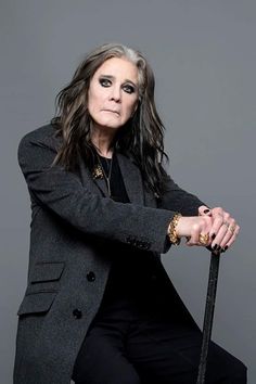 an older woman with long hair holding a cane and posing for a photo in front of a gray background