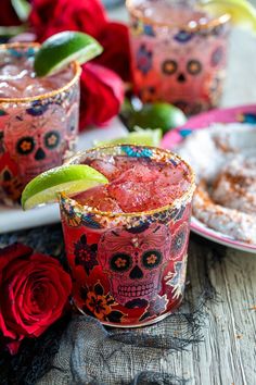 two glasses filled with colorful drinks on top of a wooden table next to red roses