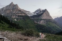 two people walking up a mountain trail in front of snow capped mountains and evergreen trees