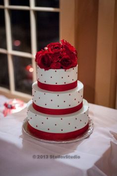 a three tiered wedding cake with red roses on top is sitting on a table