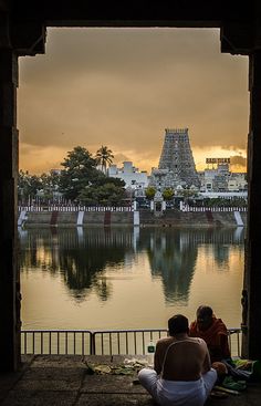 two people sitting on the edge of a lake looking out at an ornate building in the distance