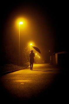 a person walking down the street at night with an umbrella in their hand and lights on