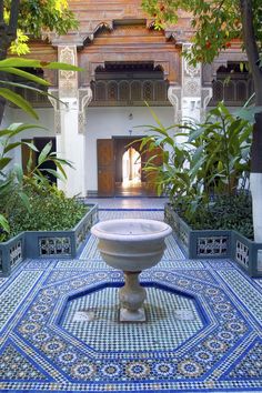 a fountain in the middle of a courtyard surrounded by plants and trees, with an archway leading to another building