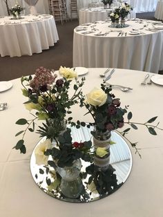 two vases with flowers are sitting on a round table in front of white clothed tables