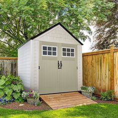 a small shed in the middle of a yard with flowers and trees around it, next to a wooden fence