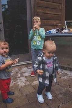 three small children standing on a brick patio eating donuts and looking at the camera