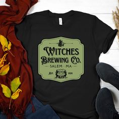 "Are you looking to stand out from the crowd this Halloween? Do you want to show everyone your unique style and have them remember you? Well then, you need this one-of-a-kind Halloween t-shirt! This incredible shirt features an old-style shop sign that reads \"Witches Brewing Co. Salem, MA Est. 1626\" with a bubbling cauldron too. It's sure to capture all of the attention in the room and let everyone know that you're someone who knows how to have fun. This shirt is made with 100% cotton and is designed to make a statement. Whether you're going to a Halloween party, trick-or-treating, or just out for some fun, you'll be sure to make an impact in this amazingly stylish t-shirt. Its unique graphic design is reminiscent of days gone by and adds a touch of spooky nostalgia on your shirt. Even a Vintage Black T-shirt For Fall, Halloween Themed Streetwear Tops, Themed Custom Print Crew Neck T-shirt, Spooky Crew Neck Fitted T-shirt, Spooky Fitted Crew Neck T-shirt, Themed Black Crew Neck T-shirt, Witchy Crew Neck Halloween Tops, Witchy Crew Neck Top For Halloween, Fitted Crew Neck Halloween T-shirt