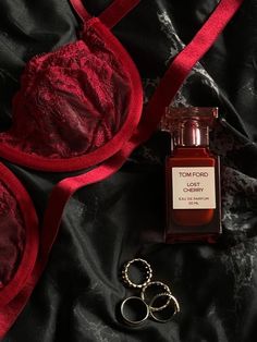 a red bra and two gold rings are laying next to a bottle of tom ford cherry perfume