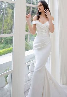 a woman in a white wedding dress standing by a window with her hand on her head