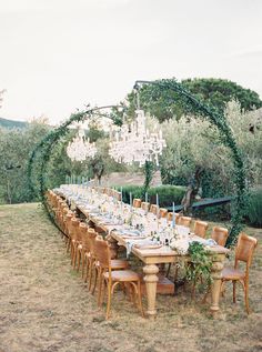 an outdoor dining table set up with chandelier and greenery in the background