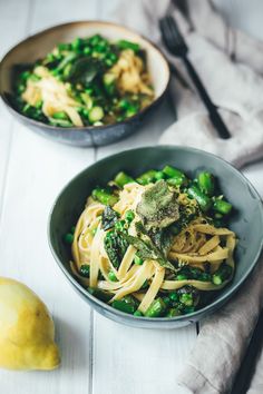 two bowls filled with pasta and vegetables on top of a white wooden table next to a lemon