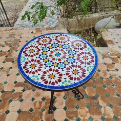 a colorful table sitting on top of a tiled floor next to a tree and bushes