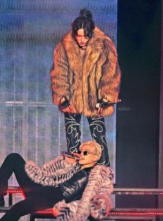 a woman standing on top of a stage next to a man in a fur coat