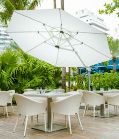 an outdoor dining area with white chairs and umbrella over the table, surrounded by palm trees