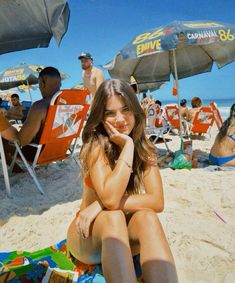 a woman sitting on top of a sandy beach next to umbrellas and people in the background