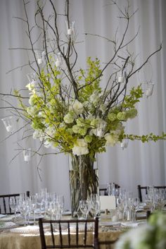 a tall vase filled with white flowers and greenery on top of a wooden table