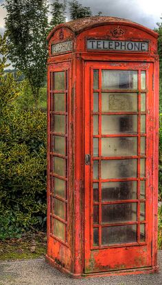 an old red phone booth sitting on the side of a road in front of some bushes