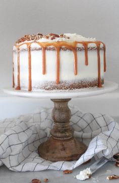 a white cake with caramel drizzle and nuts on top sitting on a pedestal