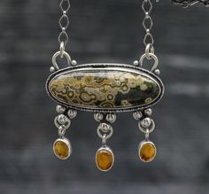 This pendant features a beautiful old stock ocean jasper. It has well defined orbs in a creamy beige with pops of yellow and a green base color. I have set it in my double bezel pendant that I like to do with three dangles of this beautiful mandarine citrine which is the perfect accent to the yellow rings in the orbs in the jasper. It pulls the color out so nicely. I have added a complimentary pattern to the back of the piece that mimics the stone. Top of the bail to bottom of the pendant it is Luxury Artisan Necklace With Patina, Bezel Pendant Ideas, Ocean Jasper Gemstone Pendant Necklace, Shiny Trinkets, Cabachon Jewelry, Iolite Necklace, Pmc Jewelry, Jewelry Closet, Silversmithing Jewelry