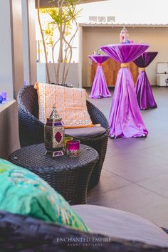 an outdoor area with wicker chairs and tables covered in purple cloths, surrounded by potted plants