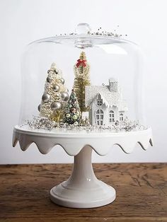 a white cake plate topped with a glass dome filled with christmas trees and houses covered in snow