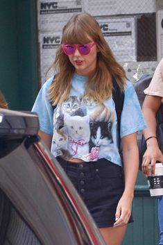 a woman with pink sunglasses and a cat t - shirt is walking past a car