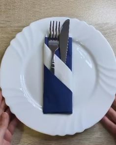 two hands holding a white plate with blue and white napkins on it, while another hand holds a fork and knife