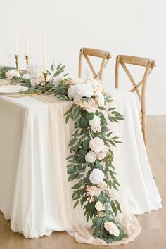 the table is set with white flowers and greenery on it, along with candles