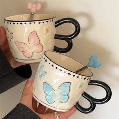 two coffee mugs with butterflies on them are being held by someone's hand