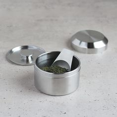 a metal container with some green stuff in it on a table next to two other containers