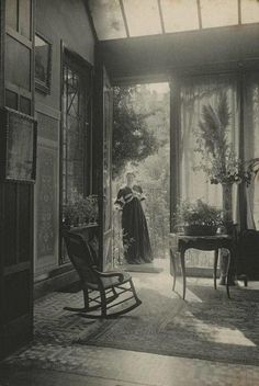 an old black and white photo of a woman standing in a room with a rocking chair