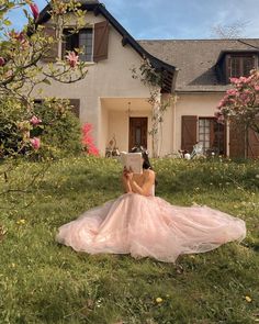 a woman in a pink dress is sitting on the grass outside her house reading a book