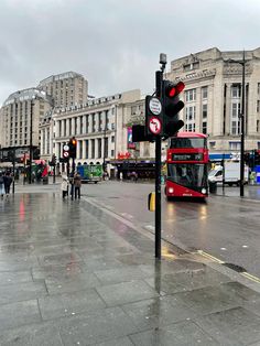 a red double decker bus driving down a street next to tall buildings and traffic lights